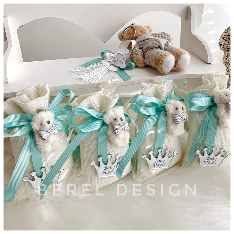 May include: Four white fabric bags with teal ribbon bows and a small white teddy bear with a silver bow tie. Each bag has a silver crown with the text "Baby Hayes" on it.
