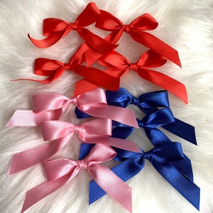 May include: A collection of satin ribbon bows in red, pink, and blue. The bows are arranged in a cluster on a white fluffy surface.