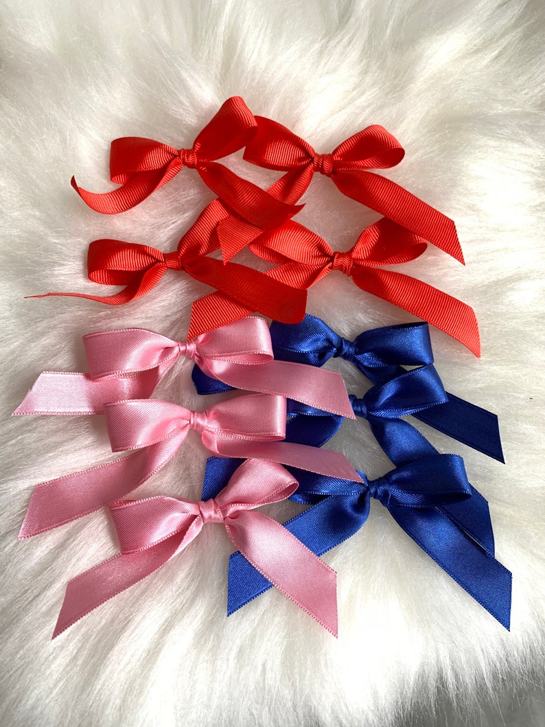 May include: A collection of satin ribbon bows in red, pink, and blue. The bows are arranged in a cluster on a white fluffy surface.