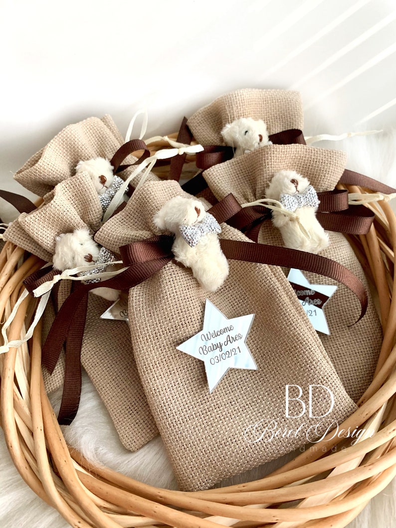 May include: A group of small burlap bags tied with brown ribbon and decorated with a white teddy bear and a silver star with the text "Welcome Baby Ares 03/02/21".