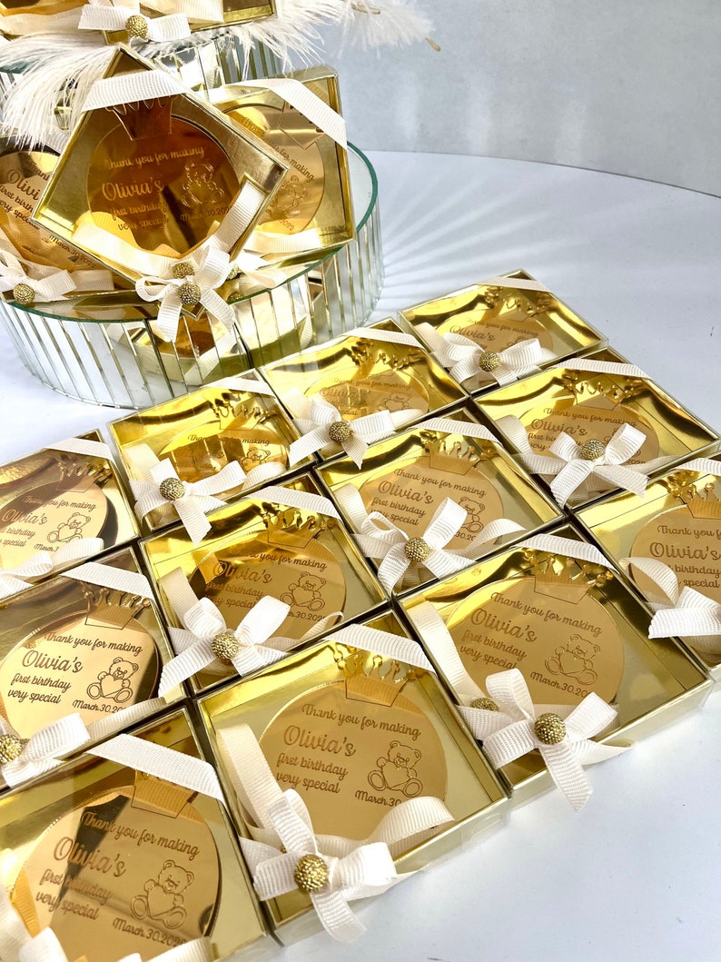 May include: Gold favor boxes with a round, gold mirror inside. The mirror has a personalized message that says "Thank you for making Olivia's first birthday very special March 30, 2020".