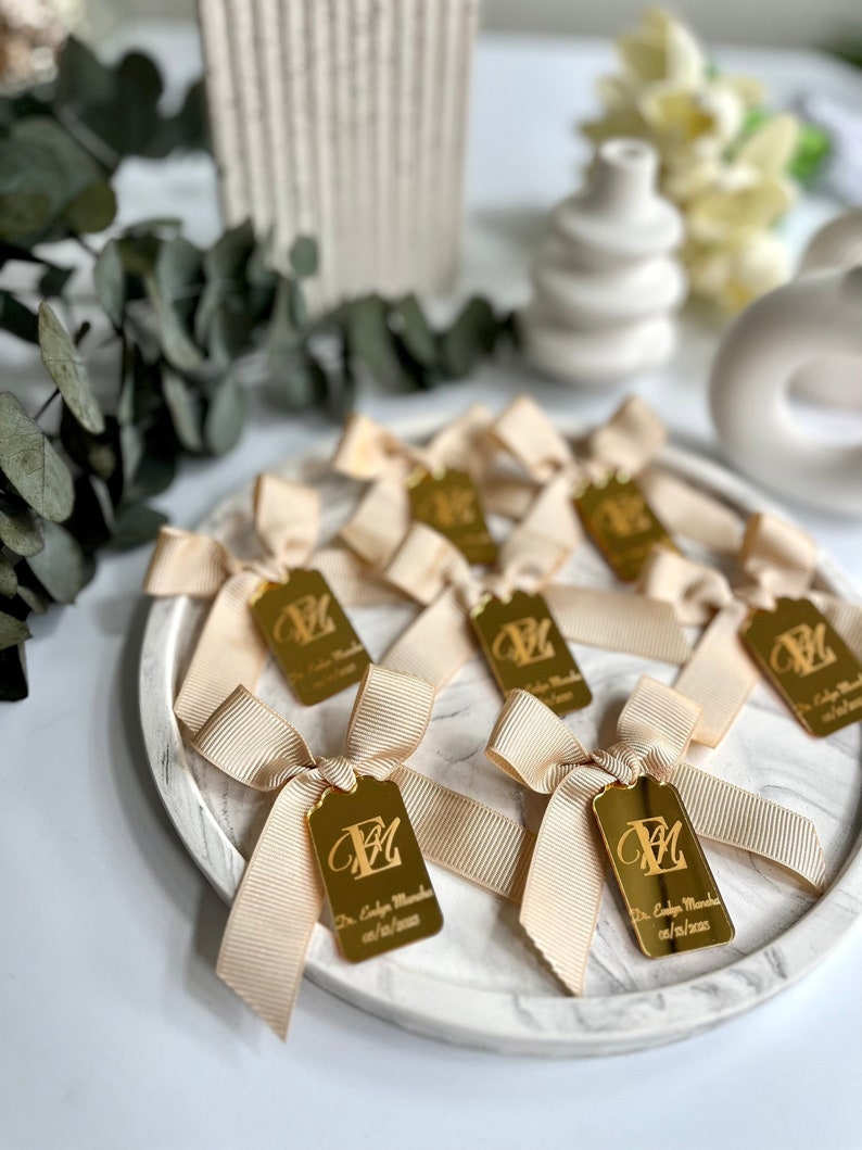 May include: A close-up of a white marble surface with gold-colored tags tied with beige ribbon bows. Each tag has a personalized inscription with the letter "M" and the text "Dr. Colby Monroe 07/16/2023".