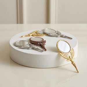 May include: A collection of four small handheld mirrors with metallic finishes in gold, silver, and copper tones. Each mirror features a circular glass surface attached to a handle shaped like a winged fairy figure with detailed wings.
