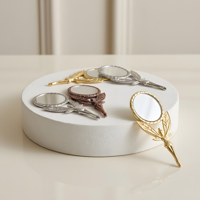 May include: A collection of four small handheld mirrors with metallic finishes in gold, silver, and copper tones. Each mirror features a circular glass surface attached to a handle shaped like a winged fairy figure with detailed wings.