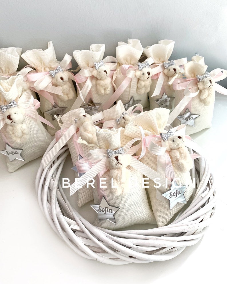 May include: White fabric gift bags with pink ribbon bows and a small teddy bear charm. Each bag has a silver star with the name "Sofia" on it.