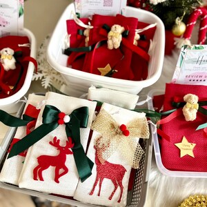 Puede incluir: Una colecci&oacute;n de peque&ntilde;as bolsas de regalo de tela roja con un dise&ntilde;o de estrella y reno. Las bolsas est&aacute;n llenas de peque&ntilde;os regalos y est&aacute;n atadas con una cinta verde y dorada. Las bolsas est&aacute;n dispuestas en una cesta blanca con un cartel que dice "&iexcl;Gracias por ser parte de nuestra familia! &iexcl;Calif&iacute;canos!"
