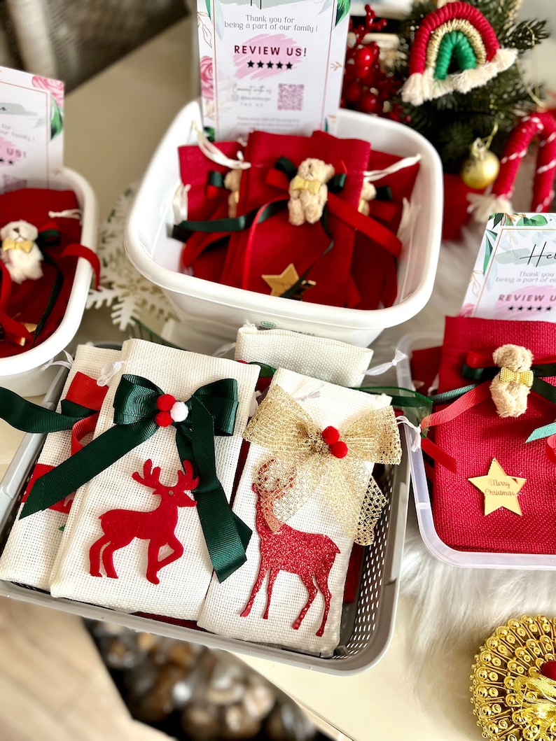 Puede incluir: Una colecci&oacute;n de peque&ntilde;as bolsas de regalo de tela roja con un dise&ntilde;o de estrella y reno. Las bolsas est&aacute;n llenas de peque&ntilde;os regalos y est&aacute;n atadas con una cinta verde y dorada. Las bolsas est&aacute;n dispuestas en una cesta blanca con un cartel que dice "&iexcl;Gracias por ser parte de nuestra familia! &iexcl;Calif&iacute;canos!"