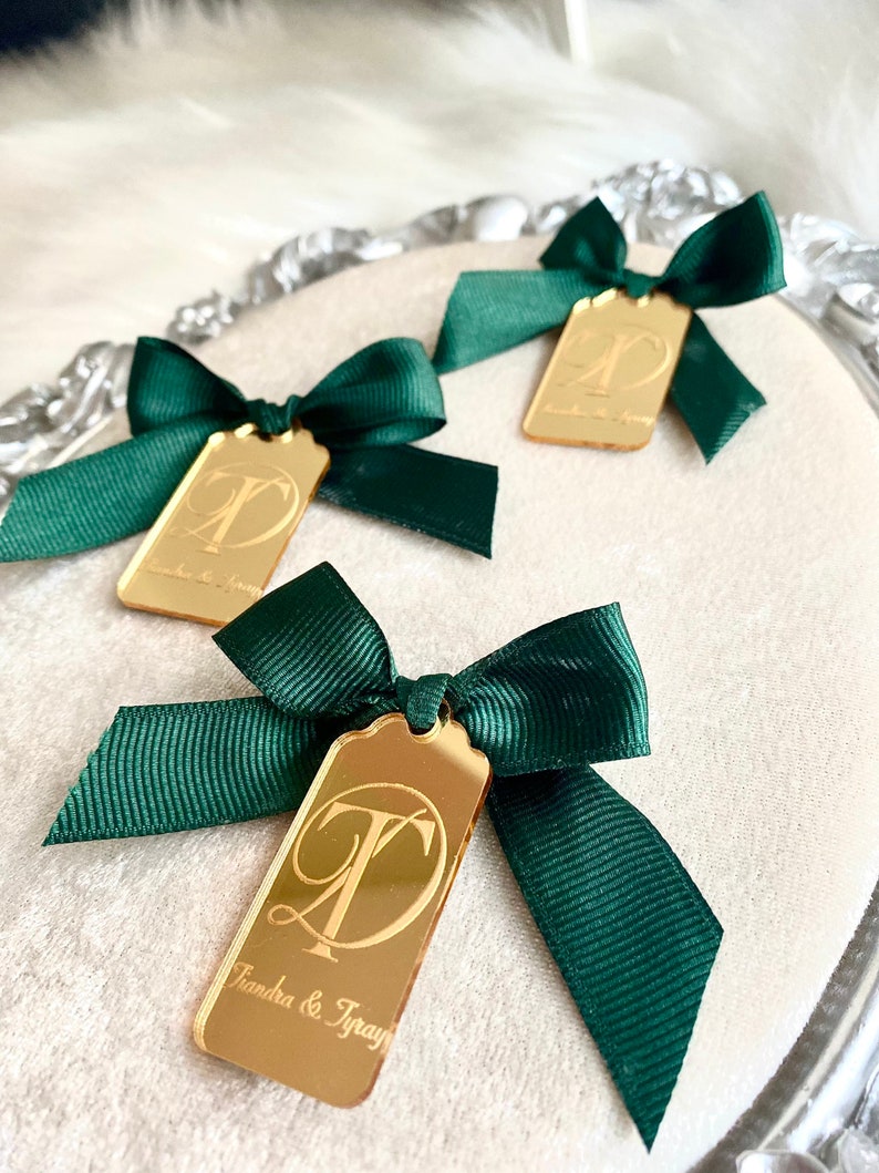 May include: Three gold mirror acrylic tags with green satin ribbon bows. The tags have a gold initial and the text "Tranda & Tray" on the bottom tag.