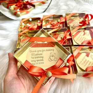 May include: Gold favor boxes with orange ribbon and a gold heart-shaped tag with the text "Thank you for making Mathias' 1st birthday very special. Feb. 22. 2022" and a small teddy bear illustration.