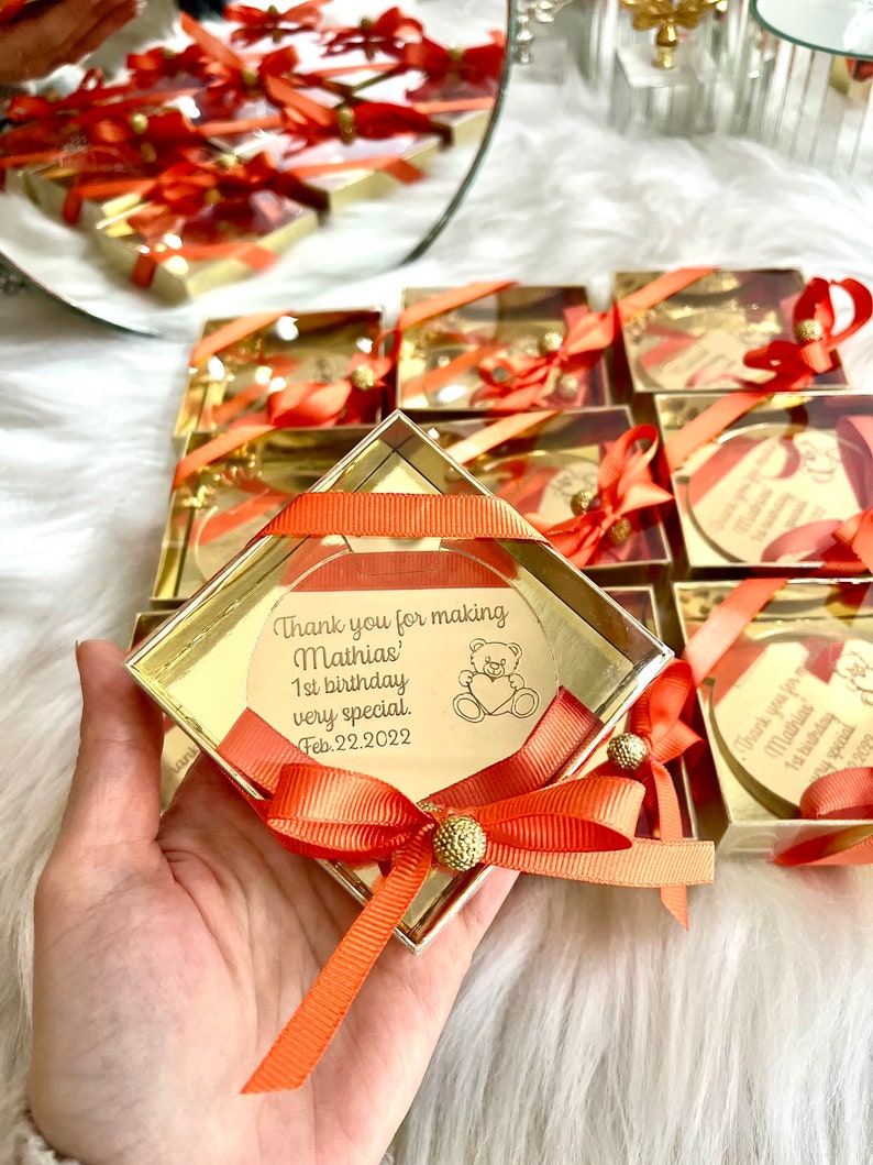 May include: Gold favor boxes with orange ribbon and a gold heart-shaped tag with the text "Thank you for making Mathias' 1st birthday very special. Feb. 22. 2022" and a small teddy bear illustration.