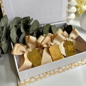 May include: A white gift box with a gold geometric pattern on the lid. Inside the box are three gold gift tags with a single initial "M" engraved on each tag. Each tag is tied with a cream-colored satin ribbon.