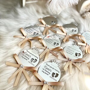 May include: A set of silver round mirror favors with a crown design and the text "Thank you for making Eleni's 1st Birthday very special 09/07/2022"  Each favor has a gold ribbon tied in a bow.