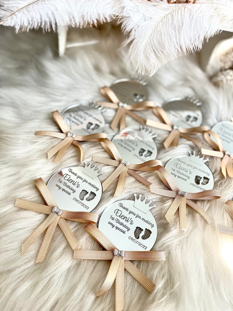 May include: A set of silver round mirror favors with a crown design and the text "Thank you for making Eleni's 1st Birthday very special 09/07/2022"  Each favor has a gold ribbon tied in a bow.