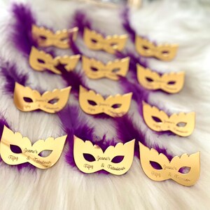Puede incluir: M&aacute;scaras doradas de carnaval con el texto "Jeane's Fifty & Fabulous" en cada m&aacute;scara. Las m&aacute;scaras est&aacute;n sobre una superficie esponjosa de color morado y blanco.