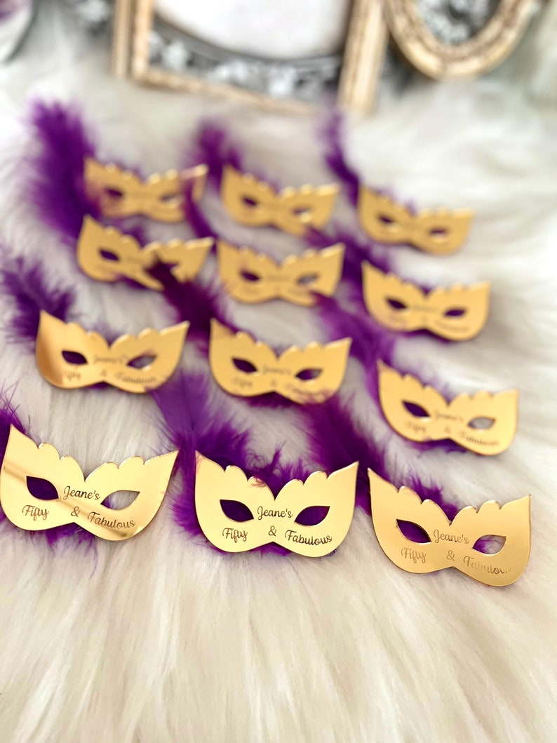 Puede incluir: M&aacute;scaras doradas de carnaval con el texto "Jeane's Fifty & Fabulous" en cada m&aacute;scara. Las m&aacute;scaras est&aacute;n sobre una superficie esponjosa de color morado y blanco.
