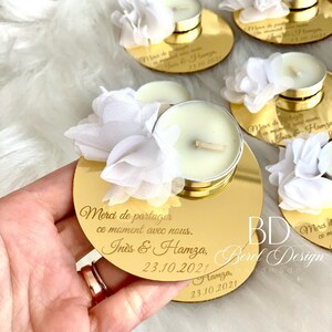 May include: Gold mirrored candle holder with a white flower embellishment. The holder has the text "Merci de partager ce moment avec nous. In&egrave;s & Hamza, 23.10.2021" engraved on it.