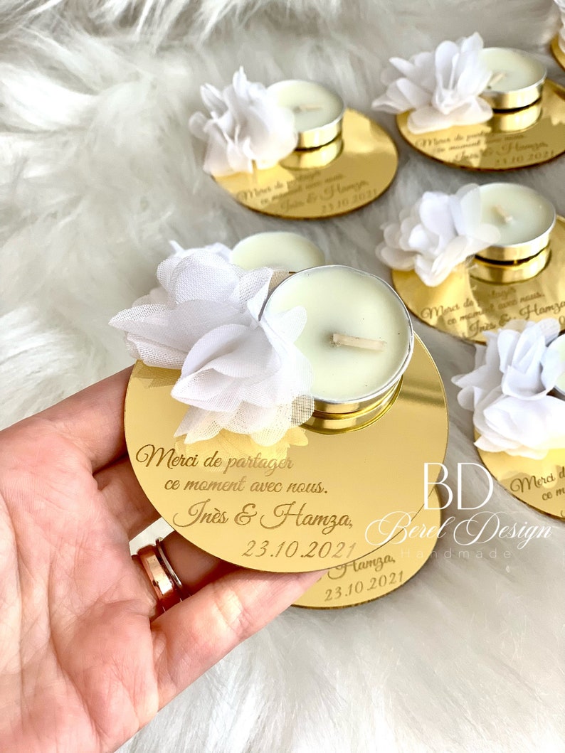 May include: Gold mirrored candle holder with a white flower embellishment. The holder has the text "Merci de partager ce moment avec nous. In&egrave;s & Hamza, 23.10.2021" engraved on it.