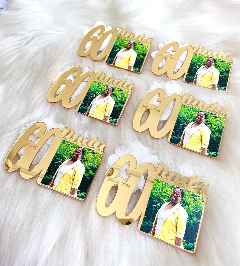 May include: Gold-colored acrylic mirror plaques with the number 60 and the name Linda. Each plaque has a small square photo of a person in a yellow shirt. The date July 15, 2022 is printed on the plaques.