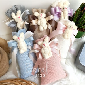 May include: Assortment of small fabric bags with bunny rabbit charms and satin ribbon bows in various colors, including pink, blue, white, brown, and purple. The bags are filled with a white substance and have the text "Berei Design" printed on one of the bags.