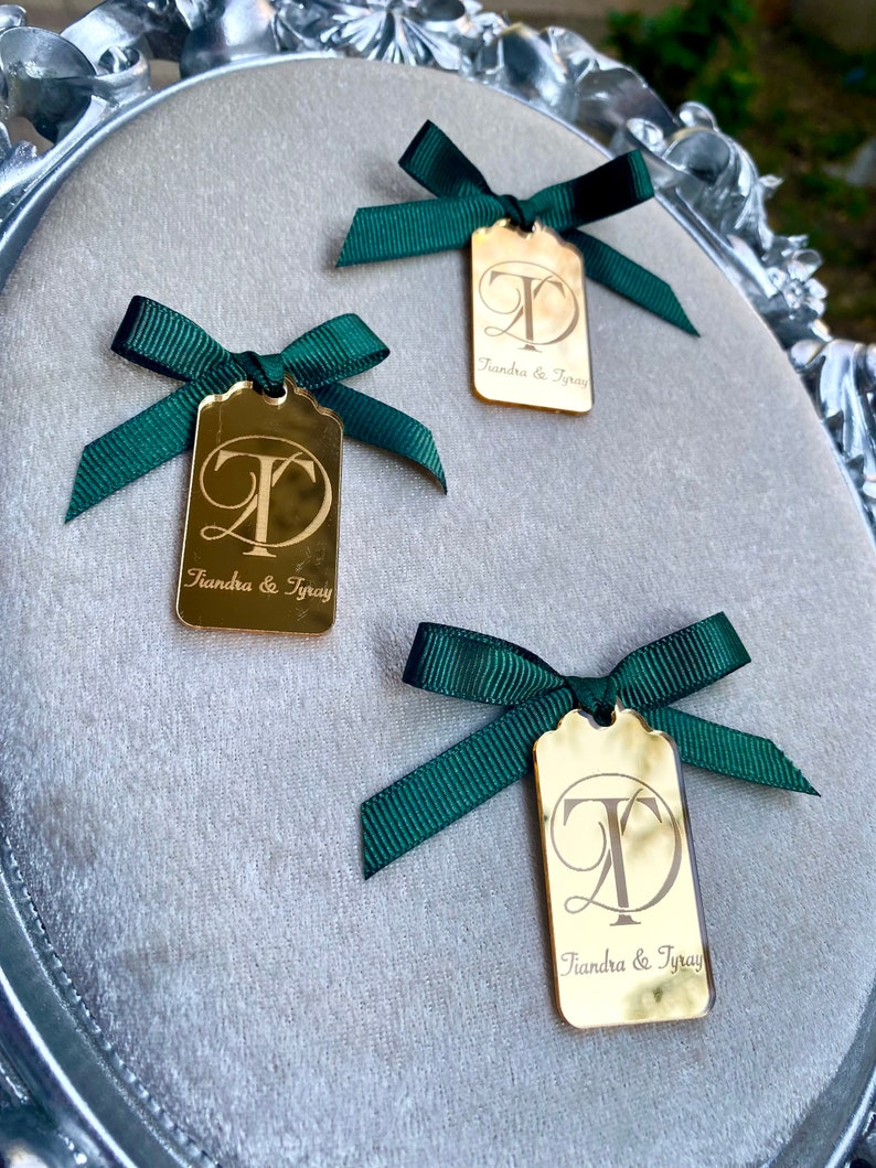 May include: Three gold mirror tags with green ribbon bows. Each tag has the letters "T" and "D" in a cursive font, with the text "Tiandra & Tyray" below.