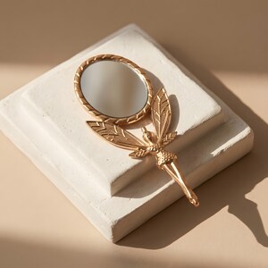 May include: A gold-toned handheld mirror featuring a circular reflective surface framed by a textured border. The handle is shaped like a winged figure with detailed leaf-like wings extending outward from the base of the mirror frame.