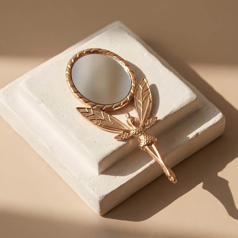 May include: A gold-toned handheld mirror featuring a circular reflective surface framed by a textured border. The handle is shaped like a winged figure with detailed leaf-like wings extending outward from the base of the mirror frame.