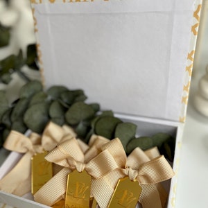 May include: A white box with a gold patterned border contains several gold-colored, rectangular, personalized favors with a bow. Each favor has a letter and the text "Dr. Cody & Mrs. [Name] 08.15.2021".