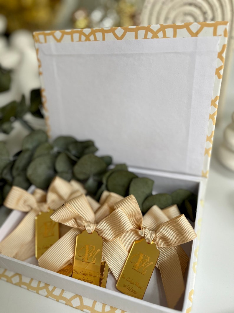 May include: A white box with a gold patterned border contains several gold-colored, rectangular, personalized favors with a bow. Each favor has a letter and the text "Dr. Cody & Mrs. [Name] 08.15.2021".