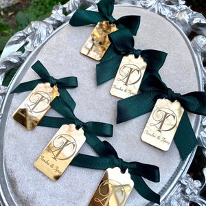 May include: Five gold mirror-like tags with a cursive "T" and the text "Tiandra & Tyray" on each. Each tag is tied with a dark green satin ribbon bow.