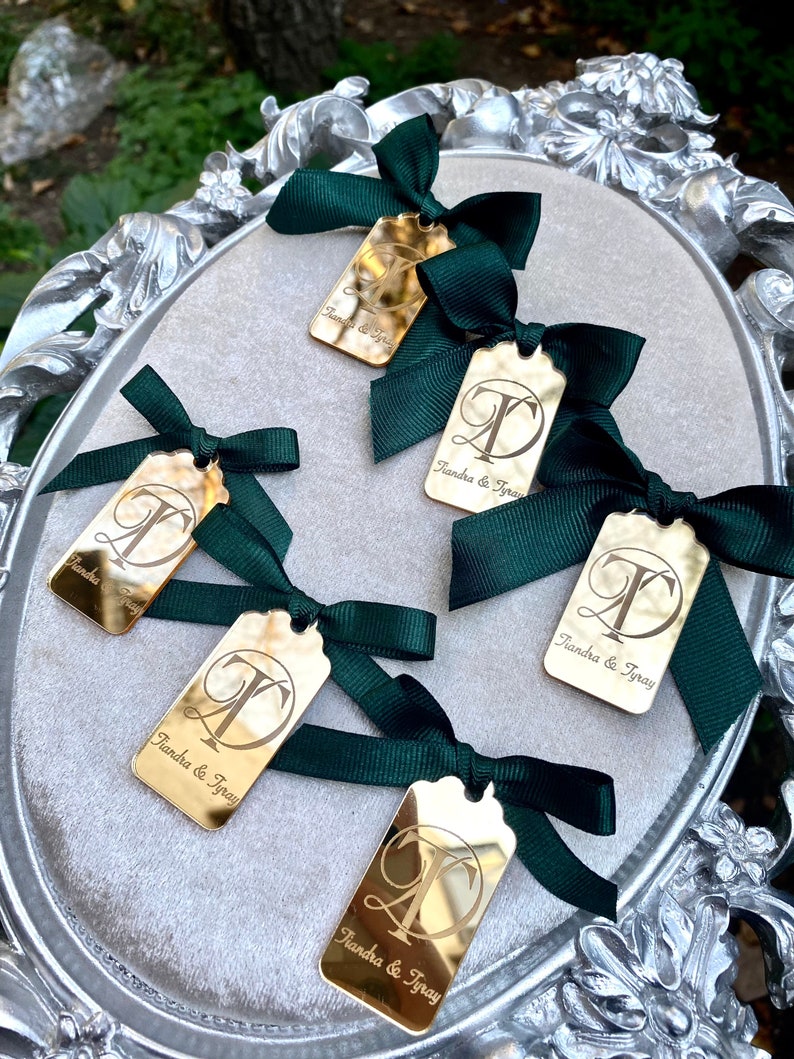 May include: Five gold mirror-like tags with a cursive "T" and the text "Tiandra & Tyray" on each. Each tag is tied with a dark green satin ribbon bow.