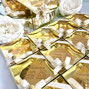 May include: Gold favor boxes with clear lids and gold mirror-like accents. Each box contains a round gold mirror with a personalized message that says "Thank you for making Olivia's first birthday very special" with a small bear graphic and the date March 30, 2022.