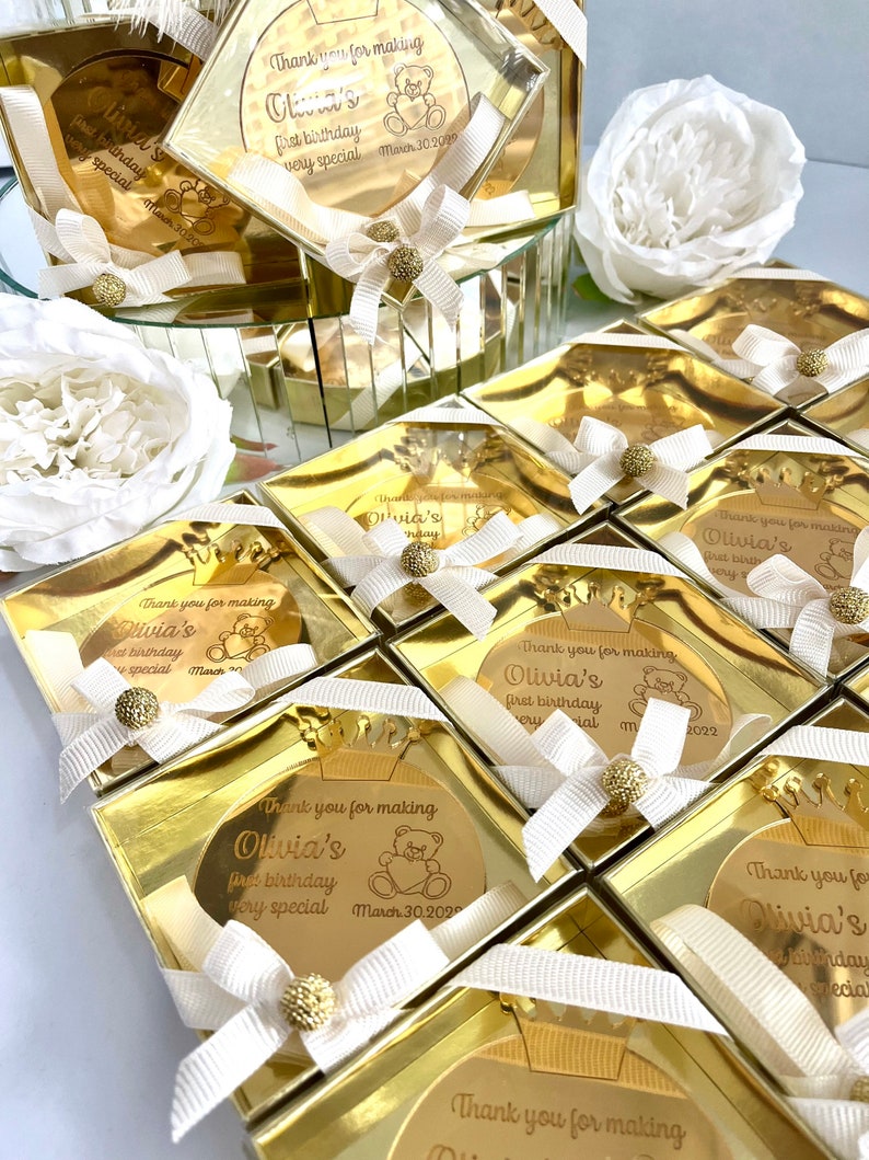 May include: Gold favor boxes with clear lids and gold mirror-like accents. Each box contains a round gold mirror with a personalized message that says "Thank you for making Olivia's first birthday very special" with a small bear graphic and the date March 30, 2022.