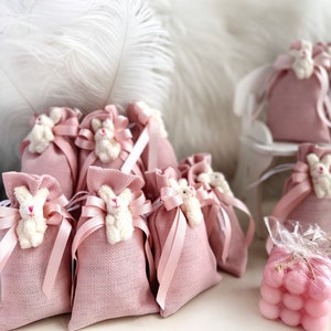 May include: Pink fabric gift bags with white bunny appliques and pink ribbon ties. The bags are arranged on a white surface with a white feather boa in the background. There are also pink bath bombs in a clear plastic bag.