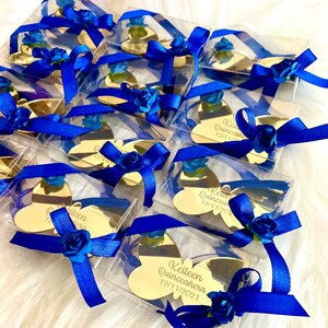 May include: Gold butterfly-shaped acrylic mirror favors with blue ribbon and blue flower accents. Each mirror has the text "Keileen Quinceanera 12/11/2021" engraved on it.