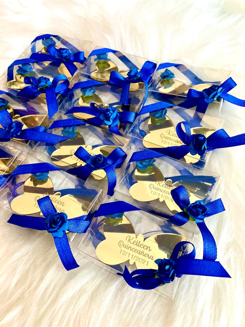 May include: Gold butterfly-shaped acrylic mirror favors with blue ribbon and blue flower accents. Each mirror has the text "Keileen Quinceanera 12/11/2021" engraved on it.