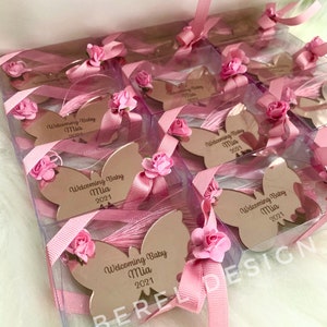 Puede incluir: Favores de espejo en forma de mariposa rosa y blanca en cajas transparentes con lazos de cinta rosa. Cada espejo tiene el texto "Welcoming Baby Mia 2021" grabado.