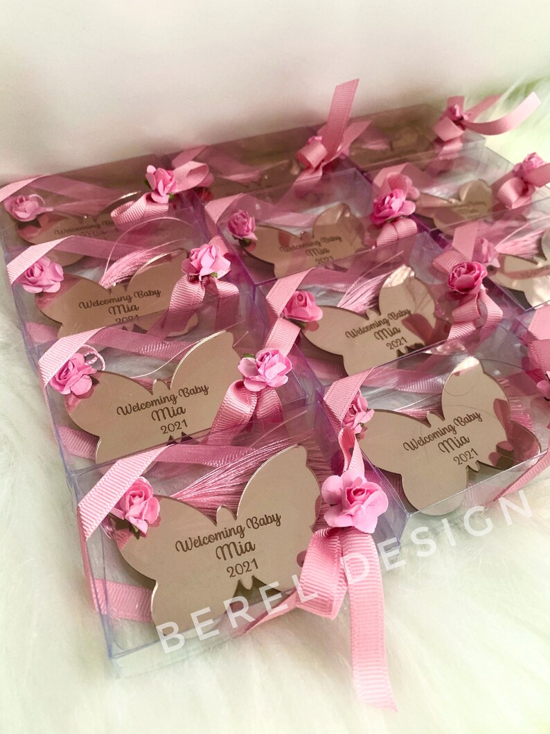 Puede incluir: Favores de espejo en forma de mariposa rosa y blanca en cajas transparentes con lazos de cinta rosa. Cada espejo tiene el texto "Welcoming Baby Mia 2021" grabado.