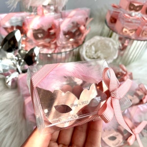 May include: A rose gold masquerade mask with the text "May 6, 2023" and "Gianna's" engraved on it. The mask is in a clear plastic box with a pink feather and pink ribbon.