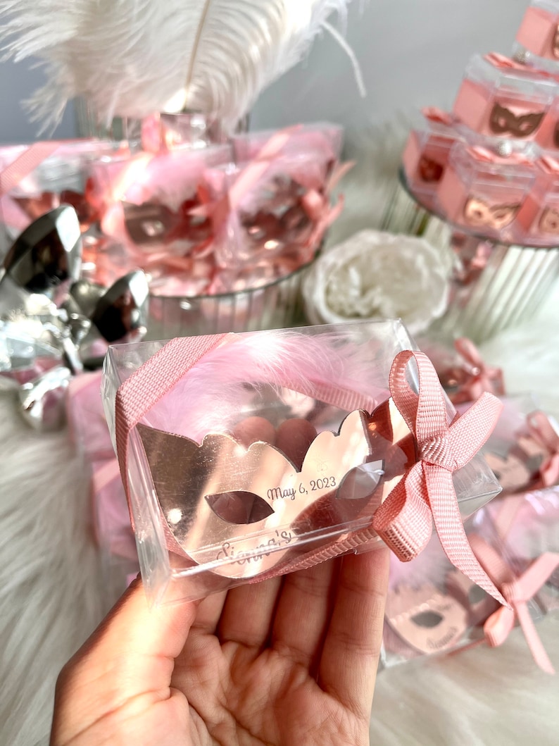 May include: A rose gold masquerade mask with the text "May 6, 2023" and "Gianna's" engraved on it. The mask is in a clear plastic box with a pink feather and pink ribbon.