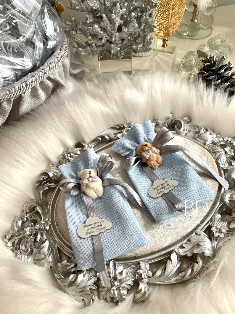 Puede incluir: Dos bolsas de regalo azul claro con cinta gris y un peque&ntilde;o amuleto de oso de peluche. Cada bolsa tiene una etiqueta plateada con el texto "&iexcl;Gracias por duchar a Baby Vinny!"