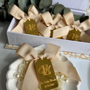 May include: Gold favor tags with a personalized monogram and the text "Dr. Evelyn Mancha 05/23/2023" tied with a beige satin ribbon and placed on a white ceramic dish with white pearl beads.