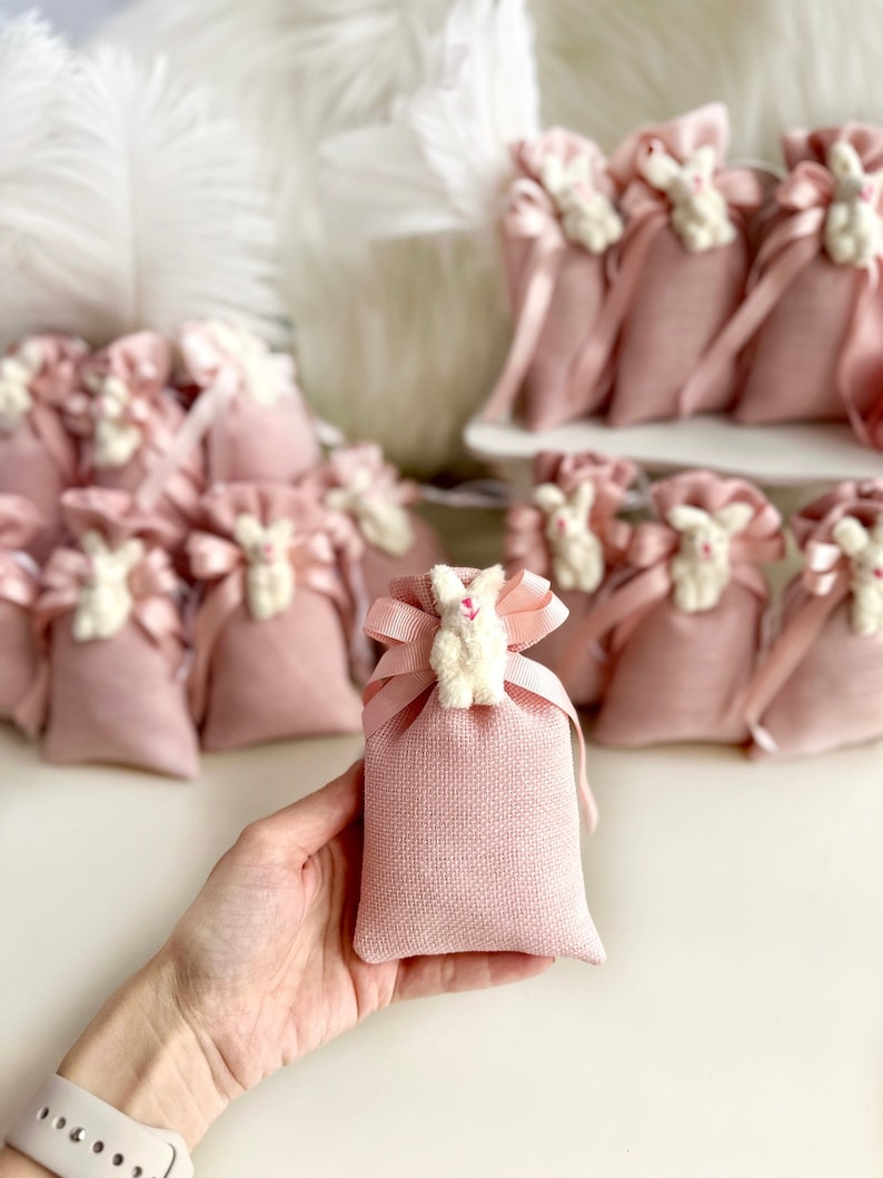May include: Pink burlap favor bags with white bunny charms. The bags are tied with a pink ribbon and are perfect for a baby shower or birthday party.