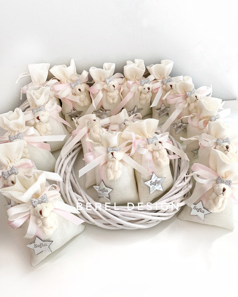 May include: A collection of white fabric gift bags with pink ribbon and a small white teddy bear charm. Each bag has a silver star with the name "Sofia" on it.