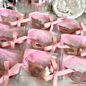 May include: Pink and gold party favours in clear boxes with pink feathers and a gold mask with the text "Vienna's May 6, 2023" on each.