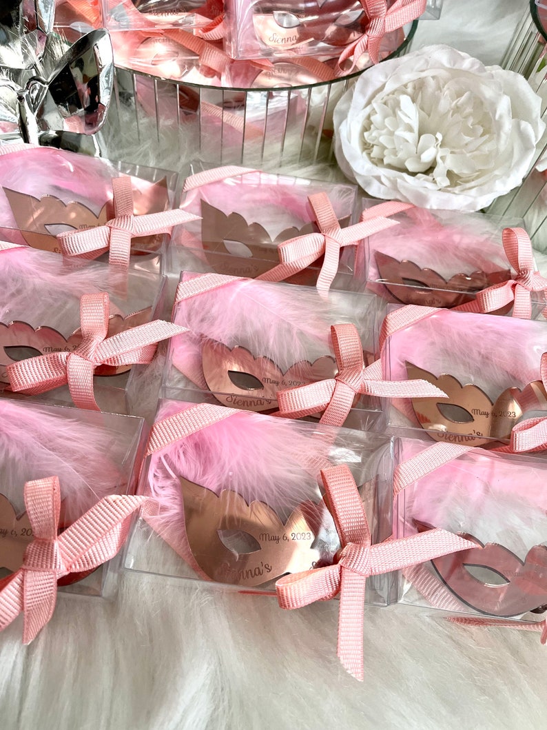 May include: Pink and gold party favours in clear boxes with pink feathers and a gold mask with the text "Vienna's May 6, 2023" on each.