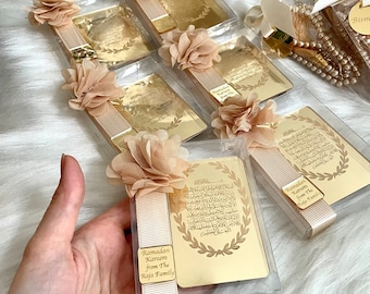 Könnte beinhalten: Goldfarbene Lesezeichen mit arabischer Kalligraphie und einem floralen Design. Die Lesezeichen sind in durchsichtigen Plastikboxen mit weißem Band und einem kleinen goldenen Etikett verpackt, auf dem "Ramadan Kareem von der Familie Raja" steht.
