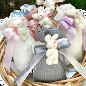 May include: A collection of small fabric bags with plush bunny toys attached. The bags are in various colors, including white, pink, gray, and purple. Each bag has a ribbon tied around it, and the bunnies have white fur.