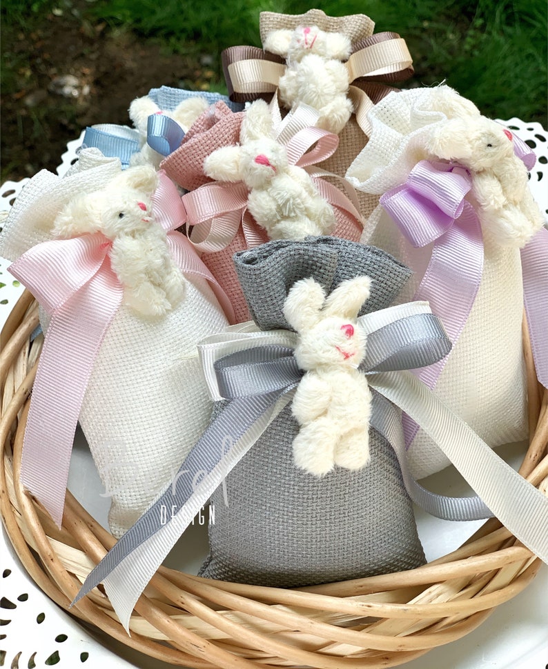 May include: A collection of small fabric bags with plush bunny toys attached. The bags are in various colors, including white, pink, gray, and purple. Each bag has a ribbon tied around it, and the bunnies have white fur.