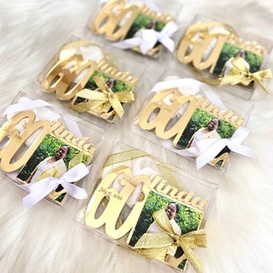 May include: Six clear boxes with gold-colored 60th birthday ornaments. Each ornament has a photo of a person and the name "Linda" on it. The boxes are tied with white and gold ribbon.