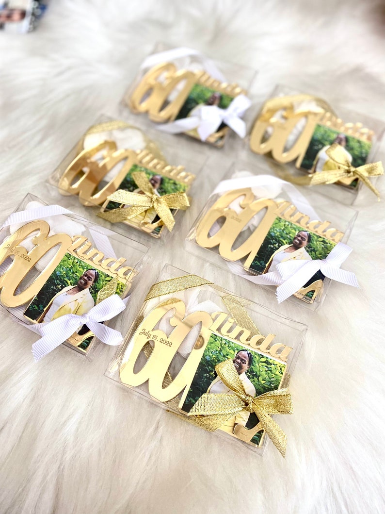 May include: Six clear boxes with gold-colored 60th birthday ornaments. Each ornament has a photo of a person and the name "Linda" on it. The boxes are tied with white and gold ribbon.